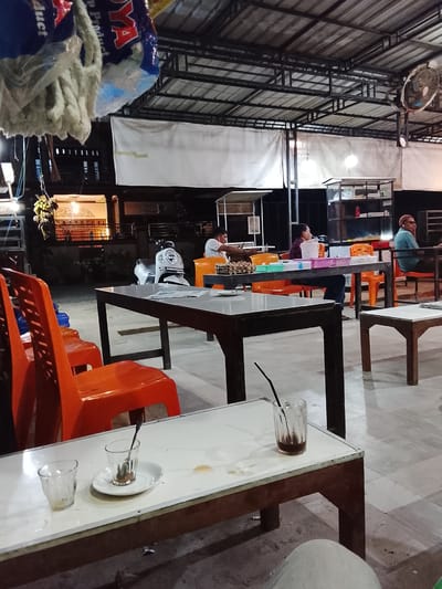The image depicts an outdoor seating area in Dayahlhoksukon Barat, Indonesia, likely a roadside cafe or warung, at night. The space is covered with a metal frame roof. Visible are several tables and orange plastic chairs arranged in a casual manner. Three men are seated at tables further back, engaged in conversation or using electronic devices. A white scooter is parked nearby. In the foreground, a small table holds two empty glasses and a saucer with a spoon, suggesting recently finished drinks. A partially full glass with a straw also sits on another small table. The flooring is tiled. The area is illuminated by electric lights. Hanging from the roof are what appear to be cleaning supplies in bags. A building with illuminated windows and a patterned balcony is visible in the background.