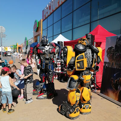 The image depicts an outdoor event taking place on a clear, sunny day in a commercial area within Трубачеевка, Russia. Two individuals are prominently featured in elaborate full-body costumes of Transformers characters: Optimus Prime, positioned on the left, rendered in metallic grey, blue, and red with flame decals; and Bumblebee, on the right, distinguished by yellow and black armor with visible wheel components integrated into the design.
A crowd, primarily composed of children of various ages and some adults, is gathered around the costumed characters on a light brown paver-tiled ground. Children are casually dressed in summer attire, with some observing the characters intently. One boy in a light blue tank top and patterned shorts holds a black spherical object. Another boy in a black and white patterned t-shirt and cargo shorts, along with a girl in a light blue top, appears to be examining an item resembling a tablet or paper. An orange balloon is visible near a stroller in the mid-ground on the left.
The background features a large, modern commercial building with a reflective glass facade and yellow paneling. Large white Cyrillic letters spelling "РЕСТОРАН" (RESTAURANT) are visible on the upper part of the building. Below, prominent green circular logos identify "Пятерочка" (Pyaterochka, a Russian supermarket chain) and "Детский мир" (Detsky Mir, a Russian children's goods store). In front of the building, a red pop-up canopy tent and a white pop-up tent are erected. A black audio speaker stands beneath the red tent, adjacent to the Bumblebee costume. To the right of Bumblebee, a large vertical banner displaying "TRANS" and other Transformers imagery is present. Overhead, the sky is clear and blue, indicating bright daylight. In the distant background, a street with vehicles, including a white car and a dark motorcycle, is visible.