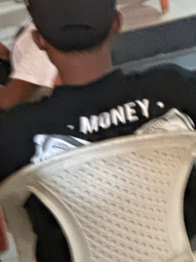 An overhead-angle image depicts the upper torso and head of an individual seated in Iriebe, Nigeria. The subject wears a dark cap and a black long-sleeved shirt with the word "MONEY" printed in white capital letters on the upper back, flanked by two small white circular marks. The individual's neck shows dark skin. Directly in front of the subject, partially obscuring the lower back, is the perforated backrest of a light-colored plastic chair. To the left, a portion of another person wearing a light-colored short-sleeved garment is visible. The background includes indistinct dark and orange surfaces. No specific actions or interactions are discernible, and the lighting suggests an indoor or evening setting.