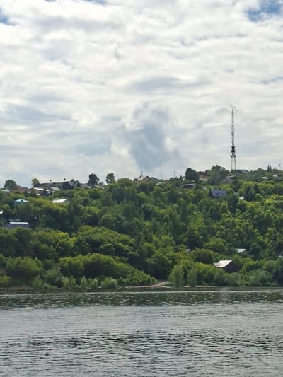 The image shows a view across a body of water towards a densely wooded hillside dotted with houses. The water in the foreground is rippled, reflecting the overcast sky. The hillside is covered in a variety of green trees, with some houses peeking through the foliage. On the crest of the hill, a tall transmission tower stands against the cloudy sky. The architecture of the houses appears to be simple, with pitched roofs. A small, dark-colored shed or outbuilding with a light-colored roof is visible nestled among the trees on the hillside. The overall scene suggests a rural or semi-rural environment. This image was captured in Verkhny Uslon, Russia.