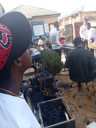 This image depicts an outdoor musical performance or rehearsal in Jebbu Miango, Nigeria. The perspective is from behind and slightly above the right shoulder of a person wearing a black baseball cap with red and white patterning, a white t-shirt, and a gold chain.
In the immediate foreground, a stack of black audio rack-mount equipment is visible on the ground, entangled with numerous multicolored cables. An open black flight case, filled with coiled blue and black cables, rests next to the equipment stack.
In the midground, a musical ensemble is active. A man in a white turtleneck sweater stands, facing right, positioned behind a black electronic keyboard on a stand. To his right, a person wearing a dark jacket is seated with their back to the camera, next to a drum cymbal on a stand, indicating a drum set. A large dark speaker on a stand is positioned behind the keyboardist. To the left of the keyboardist in white, another person is seated, operating a second electronic keyboard. Further in the background, another individual is seen playing an electric guitar.
The background features a building with a green corrugated metal roof and a blue sign with white text affixed to its wall. Adjacent to this is a multi-story building with orange-brown walls and balconies. Several individuals are visible observing the scene in the background. The ground is unpaved, likely dirt or compacted earth, with various cables running across it.