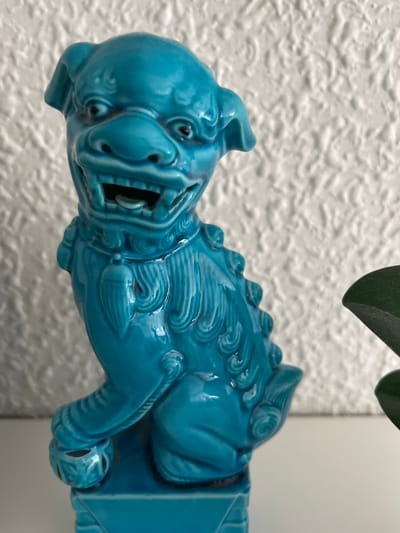 This image showcases a close-up of a vibrant turquoise ceramic guardian lion figurine. The figurine is depicted in profile, with its head facing the left side of the frame. The lion has a fierce expression, with its mouth open in a snarl, revealing sharp teeth. Its eyes are small and dark, and its face is intricately detailed with wrinkles and creases, giving it a somewhat menacing appearance. The body of the lion is adorned with stylized waves and curls, suggesting movement or a flowing mane. Small, rounded spikes run along its back. The figurine appears to be supported by a darker, rectangular base or structure, only partially visible.
The background consists of a textured white wall, possibly stucco or a similar material, which provides a neutral backdrop that makes the bright color of the figurine stand out. In the upper right corner, a hint of green foliage is visible, suggesting the figurine might be part of a larger display or placed near a plant.
The lighting in the image is bright, suggesting it was taken in daylight. There are no people or explicit activities depicted. The focus is entirely on the detailed craftsmanship of the ceramic lion. The location context provided is Torrent, Spain, which doesn't offer specific visual cues within the image itself, but adds a geographical context to the object's origin or display. The overall mood conveyed by the figurine is one of strength and perhaps a bit of playful ferocity, due to its stylized and bright execution. No text is visible in the image.
