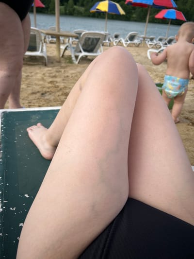 The image shows a first-person perspective of a person relaxing on a lounge chair on a sandy beach, with a body of water in the background. In the foreground, the person's legs and feet are visible, with the legs crossed. A person in the background wearing a diaper is walking away from the camera. Several beach chairs and umbrellas are set up on the sand. The general environment appears to be a recreational area with a lake or river. The specific location in Yubileyny, Russia, is not explicitly identifiable from the visual information provided in the image.