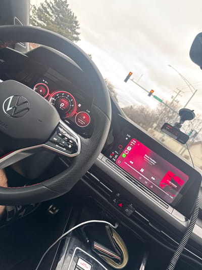 The image depicts the interior perspective of a modern Volkswagen GTI vehicle, stationary at an intersection in Bloomingdale, United States of America. The vehicle is facing an intersection with two traffic lights, both illuminated red. A green street sign, partially visible, indicates "SCHM... RD," consistent with Schmale Road.
The time displayed on the digital instrument cluster and the infotainment system is 3:05. The external temperature is 35 degrees Fahrenheit, suggesting cool or cold weather. The sky is overcast.
Inside the vehicle, a black multi-function steering wheel with red stitching and a central VW logo is prominent. The digital instrument cluster shows "GTI," a speed of 0 MPH, 17,896 miles on the odometer, and an average fuel economy of 25.6 MPG. The infotainment system displays Apple CarPlay, actively playing the song "Black Panther" by Nle Choppa & Bp FAL, with the volume at 75%. A dashcam is mounted on the windshield via a suction cup. A driver's left arm is partially visible on the lower-left side of the frame. A white charging cable is connected to a device in the center console area, near the engine start/stop button and a hazard light button.
Outside the vehicle, through the windshield, bare deciduous trees and some evergreen foliage are visible, indicative of a dormant season. Utility poles and overhead wires are also present.