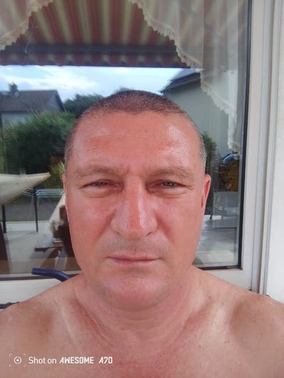 A portrait taken from a low angle captures a fair-skinned man from the shoulders up, standing in front of a window. He has short, dark hair, thinning on top, and a weathered complexion with visible sun damage and red undertones. His eyes are open and focused forward, with faint wrinkles around them. His mouth is closed, and he appears to be clean-shaven.

The background shows a view through a window, revealing a suburban setting. A pitched roof house with a chimney is visible to the left, partially obscured by green foliage. A patio table and chairs are set up outside. The window is framed by a striped awning and lace curtains. The overall impression is that of a candid self-portrait taken outdoors in a residential area of Lohmar, Germany.