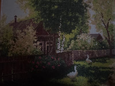 The image is a close-up, slightly blurry, photograph of a diamond painting. The painting depicts a rural scene with a red house featuring a chimney, surrounded by trees and bushes. In the foreground, there are two white geese, one to the right and one to the left of center. Pink flowers are visible in a garden area. A dark, textured fence runs horizontally across the lower part of the painting. The sky is a muted blue. The overall impression is of a tranquil countryside setting.