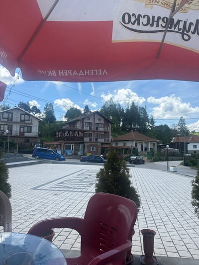 The image, taken from a slightly elevated perspective, depicts an outdoor cafe setting in a village or small town, likely in Cherna Mesta, Bulgaria. In the foreground on the left, a large red and white umbrella, possibly for a cafe or restaurant, dominates the view. Visible on the umbrella, in stylized black Cyrillic script on the white section, are the words "ВЕЛИКО ТЪРНОВО" (Veliko Tarnovo) and partially "ЗЛАТЕН ВКУС" (Golden Taste). On the red section, white Cyrillic text, reversed due to perspective, also includes "ВКУС" (Taste) and parts of "ЗЛАТЕН" (Golden), suggesting an advertisement for a beverage, likely beer.
To the right and bottom of the umbrella are parts of red and wicker chairs, and a glass-topped table, along with a brown ash urn on a stand.
Beyond the cafe seating, a light grey paved area, resembling a sidewalk or square with rectangular tiles, stretches into the background. Several multi-story buildings, some with balconies and traditional pitched roofs, line what appears to be a street or pathway. These buildings vary in architectural style, some appearing residential and others potentially small businesses. Parked vehicles, including a blue car and a black car, are visible on the street. Lush green trees and bushes are scattered throughout the scene and on a hillside in the background.
The sky is bright blue with scattered white clouds, indicating a clear, sunny day, likely midday or early afternoon, given the strong shadows cast on the paved ground. No people are visible, and the scene appears peaceful and quiet.