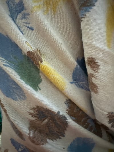 The image is a close-up, overhead shot of a cream-colored fabric with a watercolor-like pattern of abstract brushstrokes in shades of blue, green, yellow, and brown. The fabric is draped and creased, creating shadows and highlighting the texture of the material. The pattern resembles a stylized floral or feather motif. There are no people or identifiable landmarks visible. The location of this image within Shamalgan, Kazakhstan is not provided by the image itself.