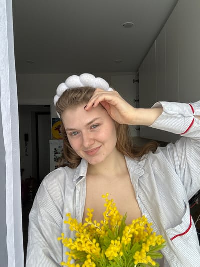 A young adult female is depicted indoors in Nikol'skii, Russia, captured from the chest up. She possesses fair skin, light brown hair, and visible freckles. On her head, she wears a white, segmented headband. Her attire consists of a light-colored, striped collared shirt, unbuttoned, revealing her chest, with red trim visible on the interior of the cuffs and a pocket. Her left hand, with dark nail polish, is raised, fingers resting near her hairline on her forehead. She holds a bouquet of yellow mimosa flowers with green foliage centrally in front of her chest. The subject is looking directly at the camera with a slight smile. The background on the left features a sheer white curtain, an indistinct dark area with a wall-mounted object, a partially visible appliance (possibly a refrigerator) adorned with items, and a yellow circular artwork. The background on the right displays a series of white, flat-panel cabinets or closet doors. The ceiling is white and includes two visible recessed lights.