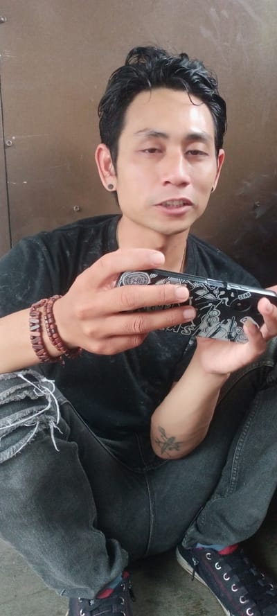 An adult male is observed sitting on a dark grey ground surface against a light brown, possibly metal, wall in Cilampeni, Indonesia. He is positioned facing the camera, with his mouth slightly open, suggesting he is speaking. His black, wavy hair is combed back, and he has dark circular ear gauges in both ears. He wears a faded black short-sleeved t-shirt and dark grey ripped jeans with visible white threads. Multiple brown wooden bead bracelets are on his left wrist, and a tattoo, possibly an anchor, is visible on his inner right forearm. He wears dark sneakers with red accents and white eyelets, paired with red socks. He holds a black smartphone horizontally with both hands, its back visible, featuring a decorative case with white illustrations and text, and a camera module in the upper left.