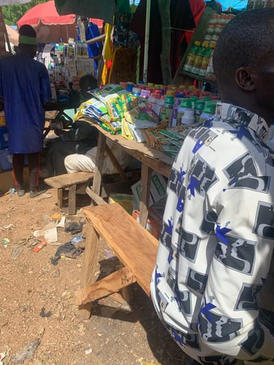 This media file depicts a bustling outdoor market scene in Nigeria. The subjects are primarily two individuals, one wearing a white shirt with a bold black and blue graphic print, and another in a blue robe with a green headband. The man in the white shirt is partially visible, his head and upper body captured as he stands behind a wooden stall laden with various goods. The second individual, wearing the blue robe, is seen from the back, appearing to be a vendor or shopper in the foreground.
The stall itself is filled with an assortment of products, including what appear to be packaged food items, spices, or agricultural supplies, judging by the colorful labels on many of the containers and bags. Some of these items are in small bottles, while others are in larger sacks or boxes. The stall structure is constructed from rough wooden planks. The ground surrounding the stall is dirt and strewn with debris, including plastic bags and discarded materials, suggesting a rustic and perhaps informal market setting.
The overall scene is illuminated by bright sunlight, indicating it is daytime. The shadows cast by the wooden structure and the people suggest the sun is high in the sky. The atmosphere appears busy and transactional, common in a marketplace. There are no strong emotional cues evident in the limited view of the individuals.
Visible text on some of the products includes what appears to be branding and product names. For example, one package prominently displays "GLOBAL COMBI" and another visible text reads "RELISATE". The presence of these labels helps identify some of the goods being sold. The setting cues, such as the merchandise and the rough construction of the stall, strongly point to a market in a developing region, consistent with the provided location context of Nigeria.