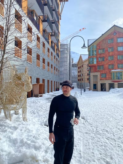 A man in a black athletic outfit stands in a snowy area, holding a cigarette and a small white cup. Buildings of various architectural styles are in the background, including a modern grey building and a brick building. A metal sculpture of a bull and a construction crane are also visible. The ground is covered in snow, and the sky is partly cloudy.