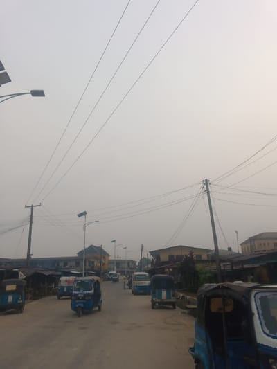 An overcast day captures a street in what appears to be a developing country. The road is lined with makeshift shops, homes, and businesses. Several blue auto-rickshaws, commonly known as "Keke Napep" in Nigeria, are visible, indicating a mode of transportation. Utility poles with numerous electrical wires crisscross overhead. Streetlights are spaced along the road, illuminating the gray atmosphere. The air seems hazy, adding to the muted color palette of the scene.