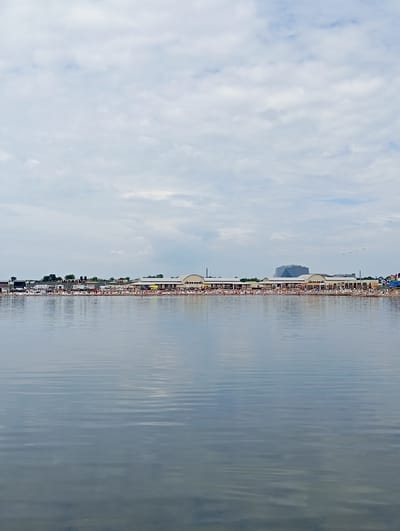 The image depicts a wide body of water in the foreground, with gentle ripples reflecting the overcast sky. In the middle ground, a long, low-lying building complex stretches horizontally along the shoreline, featuring arched openings and a beige facade. Numerous people are visible in front of this building, some seated and others standing. A variety of vehicles, including RVs and cars, are parked behind the crowd. Further back, trees and other structures, including a modern, angular building with a glass facade, rise above the horizon. The scene is located in the city of Sol-Iletsk, Russia, known for its salt lakes and tourist attractions. The overall atmosphere is one of a bustling summer day at a recreational area.