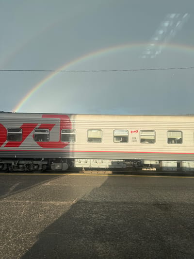 A rainbow arches across a cloudy sky above a Russian Railways train car. The train car is grey with a large red logo and several windows. The train is parked on a track next to a paved area. The scene appears to be in Kotlas, Russia.