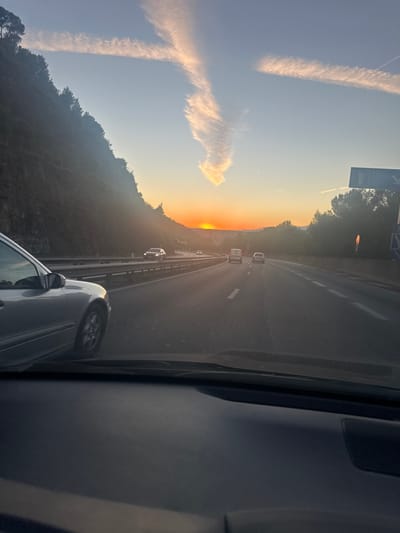 The image depicts a multi-lane highway scene at sunset, viewed from inside a vehicle looking forward through the windshield. The location is consistent with the mountainous terrain surrounding La Trinité, France, likely on the A8 motorway due to the steep, tree-covered hillside on the left.
The setting sun, a bright orange disc, is visible low on the horizon, slightly to the left of the road's center, casting strong glare and long shadows. The sky transitions from light blue at the top to a warm orange and yellow glow near the horizon. Two prominent, elongated contrails or cirrus clouds form an "X" shape high in the sky, with one arm extending downwards and broadening.
The highway features at least two lanes of traffic moving away from the viewer. White lane markings are visible on the asphalt. A metallic crash barrier runs along the left side of the road, separating it from the steep, dark green, tree-covered hillside. On the right, further trees and a partially obscured blue road sign with white text or symbols are present.
Several vehicles are on the road, traveling in the same direction. A silver sedan is partially visible in the left foreground, adjacent to the viewpoint vehicle. Further ahead, a dark sedan is in the left lane, followed by a white van or SUV in the right lane, and another light-colored sedan further ahead in the right lane. The dashboard of the viewpoint vehicle, dark and featureless, occupies the lower portion of the frame. No individuals are explicitly visible.