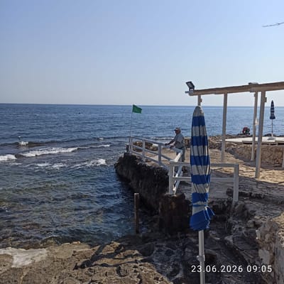 The image depicts a rocky coastline in Molfetta, Italy, with the Adriatic Sea on the left. A white railing runs along the edge of the rocky outcrop, with a man in a light-colored shirt and cap leaning on it, facing the sea. To the left of the man, a green flag flies from a pole. In the foreground, a partially closed blue and white striped umbrella stands upright. In the background, a shaded structure with white posts and a thatched roof is visible, along with another umbrella. A life preserver is also visible near the structure. The water is calm with small waves breaking on the rocks. The sky is clear and blue. A timestamp "23.06.2026 09:05" is overlaid on the bottom right corner of the image.