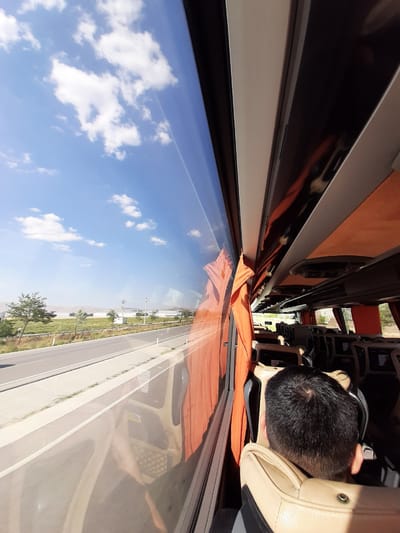 The image shows the interior of a bus traveling on a highway in the city of Karatay, Turkey, on a sunny day. The perspective is from the seat of a passenger, looking out the window. The window reflects the blue sky with scattered white clouds, as well as the orange curtains lining the interior of the bus. The highway is visible outside, with a concrete barrier and grassy embankment. In the distance, there are trees, fields, and buildings. The passenger in the foreground, a person with short black hair, is seen from the back, resting their head against the beige leather seat. The bus interior features rows of beige and orange seats, with overhead compartments and lighting fixtures.