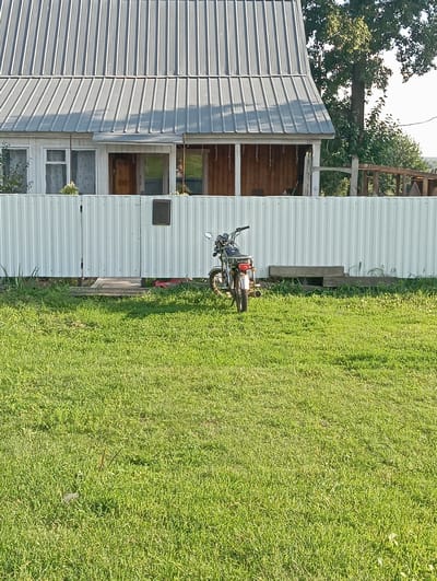In Новокопылово, Russia, a motorcycle is parked on a grassy lawn in front of a white corrugated metal fence. Behind the fence, a house with a silver metal roof and wooden-paneled porch is visible. The house has white framed windows and doors. A large tree with green leaves stands to the right of the house, and some branches of another tree are visible in the upper right corner. The sky is bright and clear. The motorcycle is dark colored with chrome accents, and the word "ALPHA" is visible on its rear. A rectangular concrete object is placed in the grass near the fence.