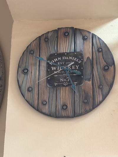 A round, wooden clock is mounted on a light-colored wall. The clock face is made of vertical wooden planks of varying shades of brown and grey, held together by what appear to be decorative screws. In the center of the clock face is a dark, rectangular metal plate with "JOHN DANIEL'S", "EST 1887", "WHISKEY", "SOUR MASH", and "No. 7" inscribed in white lettering. There are two clock hands, a shorter, wider hand that points to approximately the 11 o'clock position, and a longer, thinner hand that points to approximately the 4 o'clock position. The clock is the primary subject of the image. The background is a plain, textured wall. The scene appears to be indoors. The provided context specifies the location as Maloyaroslavets, Russia, though this information cannot be visually verified from the image alone.