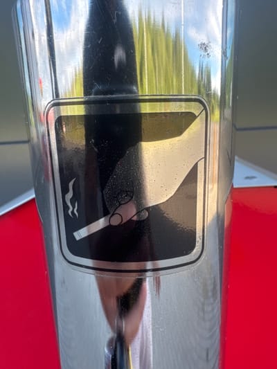 A chrome-plated cigarette disposal bin is positioned on a red surface, likely outdoors, possibly in Laagri alevik, Estonia, given the context. The bin features a black and silver symbol depicting a hand holding a cigarette and smoke, indicating a designated smoking area or disposal point. The reflective surface of the bin shows a blurred reflection of green trees and a blue sky, suggesting an exterior environment. There are minor scratches and smudges on the bin's surface, common for public fixtures.