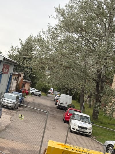 The image depicts a street scene in Příbram, Czechia. The street is lined with buildings on the left and trees on the right, with several cars parked along the side. A silver Volkswagen Golf, a blue car, a red car, and a white Skoda Fabia are visible. A white van is also parked on the street. A construction fence with a yellow sign that reads "D JOHNNY SERVIS" is in the foreground. The sky is overcast. The overall impression is of a typical urban street with some parked vehicles and signs of recent construction or maintenance.