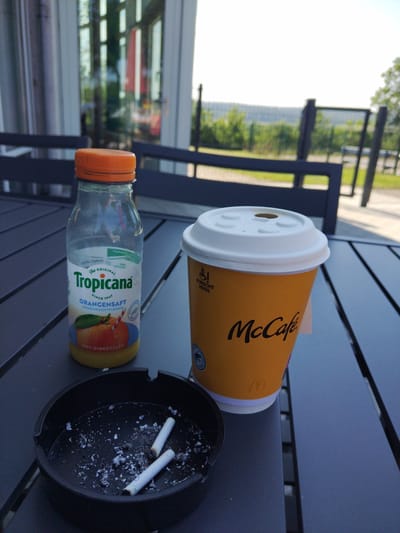 The image shows a close-up view of a dark gray outdoor table with several items on it. In the foreground, an oval-shaped black ashtray contains cigarette butts and ash. To the right of the ashtray is a bright orange McCafe coffee cup with a white lid, partially obscured by a white label. To the left of the McCafe cup is a clear glass bottle of Tropicana orange juice with an orange cap. The background shows a building with large windows, suggesting an indoor establishment, and beyond that, a slightly blurred view of greenery and sky. The overall scene appears to be an outdoor seating area, possibly at a restaurant or cafe in Oberbeisheim, Germany, given the presence of McCafe branding, a popular fast-food chain. The lighting suggests it is daytime.