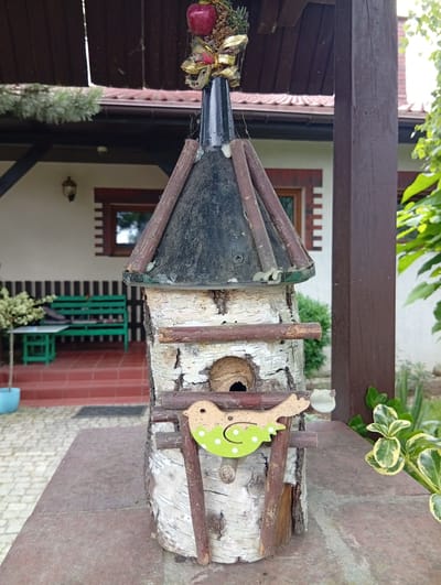 The image shows a rustic birdhouse made of birch logs and branches, topped with a dark, conical roof. A decorative red apple and festive ribbon adorn the very peak of the roof. A charming, light green wooden bird cutout with white polka dots is attached to the front of the birdhouse. The birdhouse sits on a stone surface, with a blurred background of a house, porch, green benches, and lush foliage. The scene appears to be outdoors, likely in a garden or yard, given the presence of plants and outdoor furniture. There is no visible text in the image. The lighting suggests it is daytime, with natural light illuminating the scene.