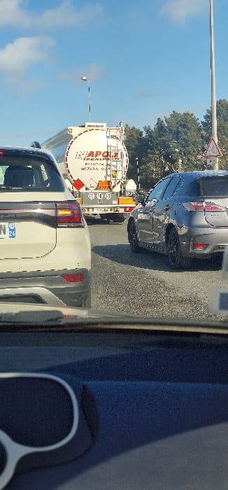The image depicts a daytime traffic scene on a road in Cenon, France. The perspective is from inside a vehicle, with the top of a dark dashboard and white-framed sunglasses visible in the immediate foreground.
In the midground, a light-colored (possibly white or light grey) SUV is directly ahead, showing its rear with a French license plate (featuring 'F' and '24'). To its right, a dark grey hatchback or compact SUV with black wheels is positioned in an adjacent lane or slightly offset.
Behind the light-colored SUV, a large white tanker truck is visible, featuring a prominent red circular logo with "MAPOJI TRANSPORTS" text. The tanker displays orange hazardous materials placards on its rear.
The background consists of a clear blue sky with scattered white clouds. A street light pole is present on the left, and a taller utility pole is on the right. Evergreen trees line the right side of the road, and a white triangular road sign with a red border is partially visible on the far right. All vehicles appear stationary in traffic.