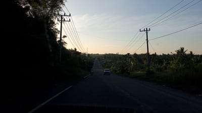 This is a photograph of a road leading through a tropical landscape near Seuneubok, Indonesia, likely at dawn or dusk due to the soft lighting. A dark, paved road extends into the distance, with a white line marking the left edge of the travel lane. On the left side of the road, a steep, heavily vegetated hill slopes upward, with bamboo and other trees casting a dark shadow. A utility pole with multiple wires stretches across the scene from the left. On the right side of the road, dense vegetation, including banana trees and palm trees, lines the shoulder. Two vehicles are visible on the road: a motorcycle with a rider in the left lane, moving away from the camera, and a dark-colored SUV in the right lane, also moving away. In the distance, the road appears to curve and wind through a village or town, with more scattered trees and buildings visible against the horizon. The sky is a pale blue with wispy clouds, and the sun is low, casting a warm glow. Another utility pole is visible on the right side of the road in the middle ground. The overall impression is one of a rural, possibly agricultural, setting within Indonesia.