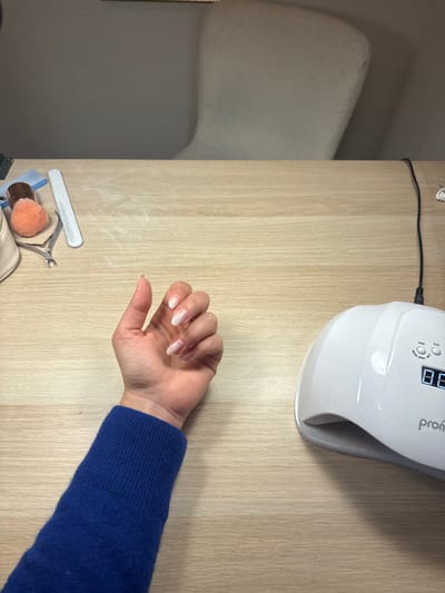 An indoor scene displays a person's left arm and hand resting on a light-colored wooden surface. The arm is covered by a royal blue, ribbed knit sweater. The visible hand has manicured fingernails, featuring a clear or light pink base with distinct white tips, consistent with a French manicure. To the left of the hand, an assortment of nail care tools is present, including a white nail file, a silver-toned cuticle nipper, a small brush with orange bristles, and a partial view of a light-colored pouch. To the right, a white UV/LED nail lamp with "prom" visible on its side sits on the table, displaying a digital reading of "88." A black power cord extends from the lamp towards the upper right corner. The background features a light-colored wall and the upper portion of an off-white, textured armchair. The specific location within Aix-en-Provence, France, is not discernible from the provided visual information.