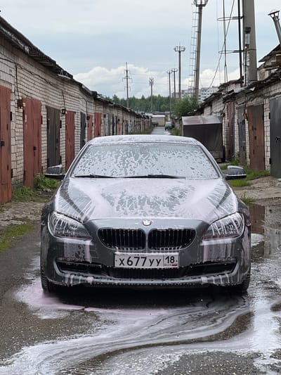 A dark gray BMW SUV is being washed, covered in pink-tinged soap suds. The car is parked on an asphalt surface, which is wet and also covered in suds, indicating it's being rinsed. The scene appears to be in a yard or service area with a row of brick garages with corrugated metal doors lining the left side of the image. The garages have a weathered look. In the background, there are power lines and tall poles, suggesting an urban or semi-urban environment. The sky is overcast with some visible clouds. The license plate of the car is partially visible, reading "X677YY 18". The overall impression is that of an outdoor car wash in a utilitarian setting.