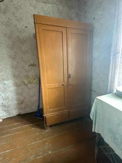 A low-angle interior shot shows a closed, light brown wooden cabinet door, likely part of built-in cabinetry, affixed to a wall covered in vintage floral wallpaper. To the right of the cabinet door, there is a portion of a wood-paneled wall. On the top right corner of the cabinet door is a piece of white machinery. Beneath the cabinet door is a light blue mop. The scene appears to be in a state of disrepair with potential water damage and aging elements visible. The location is possibly in the city of Студзёнкаўскі сельскі Савет, Belarus, based on the request context.