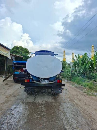 The image shows the rear of a blue and white water truck with a black chassis in Buketglumpang, Indonesia, as evidenced by the license plate. The truck is spraying water onto a dirt and concrete road. In the background, there are several buildings with yellow spires, likely part of a mosque, surrounded by green foliage and banana trees. Utility lines cross the sky, which is partially cloudy. A small, rustic building is visible to the left, partly obscured by trees.