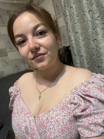 A woman is shown in a close-up selfie. She has light brown hair pulled back and is wearing a pink floral off-the-shoulder top with ruffled sleeves. She has gold hoop earrings and a gold cross necklace. The background consists of a gray couch and a patterned curtain. The image appears to be taken indoors, likely in a residential setting. There is no information in the image that suggests it was taken in the city of Zubov, Russia.