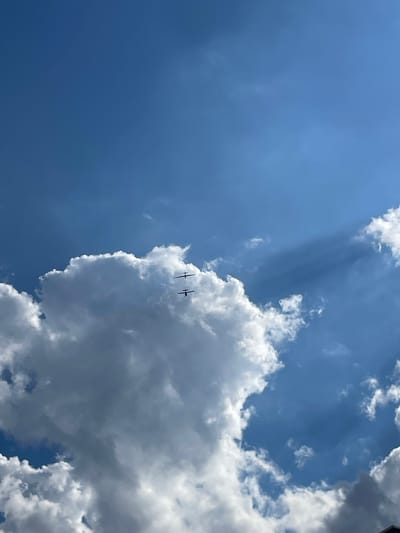 This media file captures a view of the sky on a partly cloudy day, likely during daylight hours. The sky is a vibrant blue, with several distinct clouds. One large, fluffy cloud dominates the right side of the frame, casting shadows and creating a sense of depth. There are also lighter, wispy clouds scattered across the sky, particularly in the upper left quadrant, where sunbeams are visible filtering through.
Two small, dark objects that appear to be airplanes are flying in formation near the large cloud. They are silhouetted against the bright sky, making their details difficult to discern. Their presence suggests an activity related to aviation, such as recreational flying or a demonstration.
The lighting indicates it is daytime, with the sun actively illuminating the clouds. The contrast between the bright sunlight and the shadows suggests a dynamic weather condition, moving from clear to cloudy. There are no discernible location cues or visible text within the image. The overall mood is one of openness and perhaps a touch of awe at the spectacle of flight against a dramatic sky.