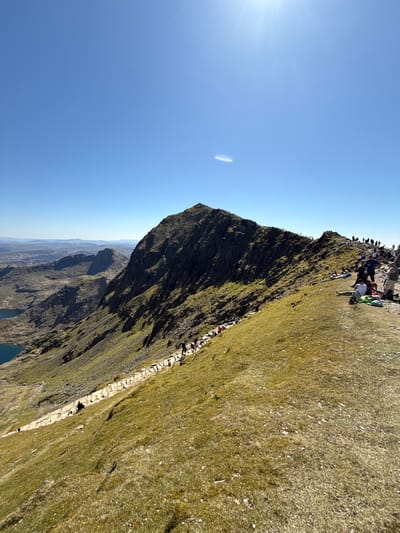 The image depicts the summit of Mount Snowdon in Beddgelert, United Kingdom, during daytime. The mountain is visible with its rocky peak and grassy slopes. Numerous people are gathered at the summit, and a stone path leads down the slope. A lake and distant mountains are seen in the background. The sky is clear blue with a bright sun.