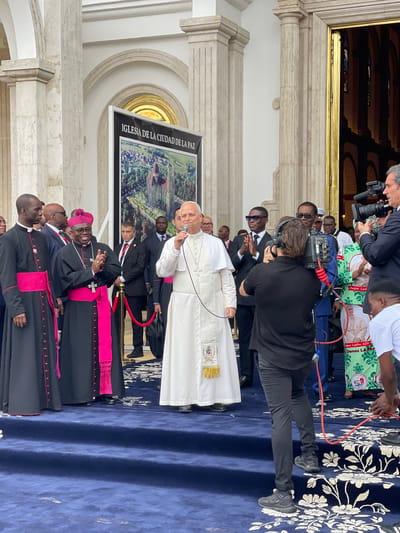 The image captures Pope Francis speaking into a microphone, likely during a visit to the Iglesia de la Ciudad de la Paz (Church of the City of Peace) in Mongomo, Equatorial Guinea. Several bishops in traditional attire, including black robes with pink trim, stand to the Pope's right. Behind the Pope, men in suits stand near a red rope barrier. A poster displays an aerial view of the church. A few members of the press, including camera operators, document the event, and another individual wears a green patterned shirt featuring an image of a past pope. The ground is covered with a blue carpet with white floral designs. The architecture of the church entrance is visible in the background.