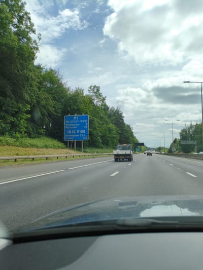 The image shows a view from inside a vehicle traveling on a highway. A blue motorway sign indicates "M5 THE SOUTH WEST", with destinations Bromsgrove (9 miles) and Worcester (23 miles), and "(M42, M40) Birmingham (S)" (123 miles). The road is a multi-lane highway with green trees and vegetation lining the left side. There are other vehicles on the road, including a flatbed truck. The sky is partly cloudy. The location is the M5 motorway, which passes near Dudley, United Kingdom.