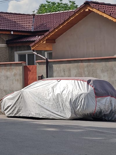 The image depicts an outdoor scene in Tsarev Brod, Bulgaria, under bright daylight. A vehicle, completely covered by a two-tone protective cover, is parked on a dark gray asphalt road in the midground. The car cover is primarily silver with a wrinkled texture, featuring a dark gray upper section and thin red piping along its seams and edges; a single dark loop is visible on the top front of the cover.
Behind the covered car, a low, light gray concrete or stucco wall topped with terracotta-colored coping separates the street from a property. Integrated into this wall on the left is a reddish-brown metal gate with a grid-like embossed pattern. To the right of the gate, a dark brown metal mailbox is affixed to the wall, and a dark brown downspout runs vertically along the wall, connecting to a gutter above. A distinct, elongated human-like shadow is cast onto the wall and gate from the left.
In the background, two residential structures are visible. The prominent house on the right features a light brown stucco facade and a roof constructed from dark reddish-brown corrugated metal tiles. Its eaves are finished with exposed light brown wood paneling and decorative trim. A second, partially obscured structure to the left also has a similar dark reddish-brown roof and a white-framed window set into a light-colored wall. Green tree foliage rises above the rooftops, and the sky is partly cloudy with white clouds against a blue background. No individuals are visible in the scene.