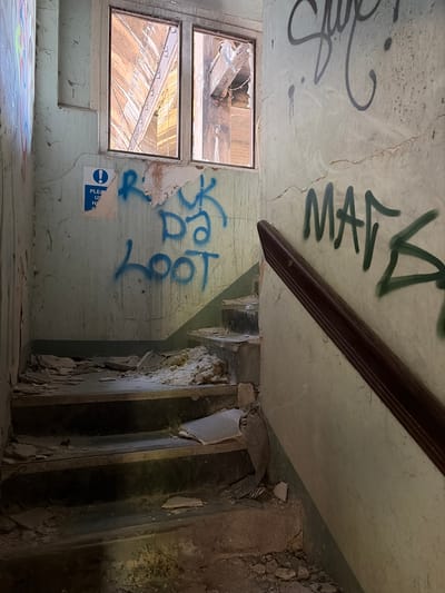 The image shows the interior of a derelict building, likely located in Mid Suffolk, United Kingdom, although this cannot be forensically confirmed. The room has two broken windows, graffiti on the walls including the word "MATA", and a damaged doorway. The floor is littered with debris. There are no people visible.