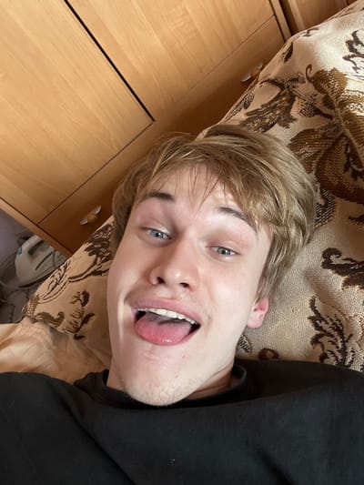 The image shows a close-up, overhead view of a young Caucasian male lying on a patterned textile, likely a bedspread. He has light brown hair and blue eyes, and he is sticking his tongue out with his mouth open. He is wearing a black long-sleeved shirt. In the background, there is a wooden cabinet with drawers, and an object that appears to be an iron is visible on the left side of the frame. The overall environment appears to be indoors, in a domestic setting. There is no specific indication of the image being taken in the city of Gari, Russia.