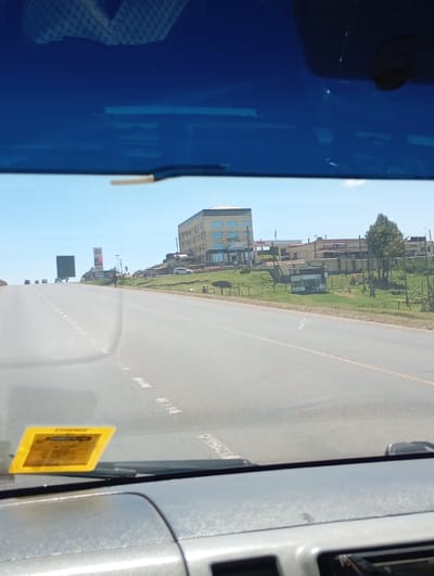 The image captures a road scene from the interior of a vehicle, looking forward through the windshield. A dark blue tint covers the top of the view, with a gray dashboard visible at the bottom. A yellow sticker, featuring text including "COVERED BY DIRECT," is affixed to the lower-left portion of the windshield.
The scene focuses on a multi-lane asphalt highway, identified as the A104, located in Naivasha East ward, Kenya. The highway features dashed white lane markings and extends into the distance, where several small vehicles are visible.
On the right side of the highway, a prominent five-story building, identified as the Rift Valley Hotel Naivasha, is situated on a raised, grassy mound. The hotel has a light-colored facade with blue-tinted windows and displays "HOTEL" on its exterior. A white car is parked near the hotel's entrance. Adjacent to and behind the hotel are additional commercial structures, including one with "TOTAL" branding and another displaying "RBS." The area around these buildings features green grass, scattered trees, and fencing. In the distant left, a large billboard structure is visible. The sky is clear and blue.