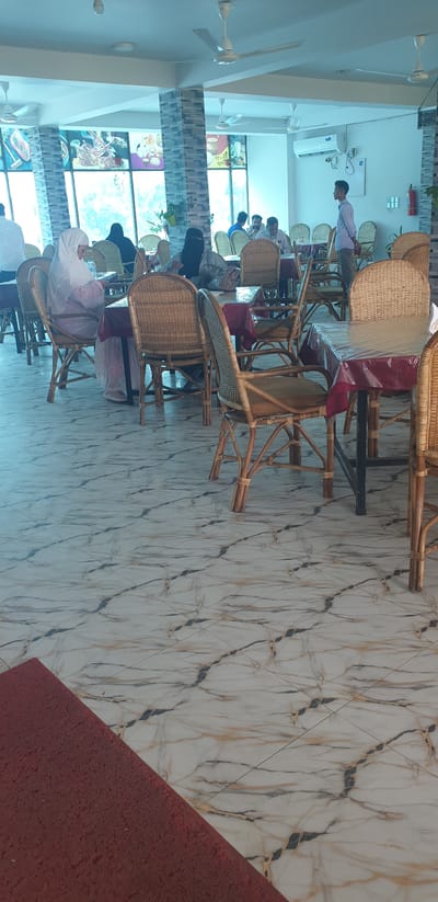 The image depicts the interior of a restaurant in Hatikumrul, Bangladesh. The restaurant features a marble-tiled floor with a red mat in the foreground. Numerous tables are covered with red tablecloths and clear plastic coverings, each surrounded by wicker chairs. Several patrons, including women in hijabs and men, are seated at the tables, engaged in dining or conversation. A young man, likely an employee, stands near the back, facing the patrons. The walls are decorated with a mosaic tile pattern, and large windows offer a view of the outdoors. Ceiling fans are visible, along with air conditioning units. The overall ambiance suggests a casual dining environment.