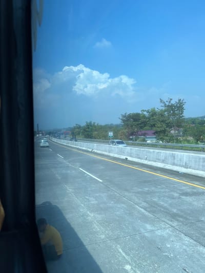 An outward-looking view from a vehicle captures a highway scene under a bright, partly cloudy sky. The highway has multiple lanes, separated by white lines and a yellow outer line. Concrete barriers line the right side of the road. Several cars are visible on the highway, some in the distance and some closer. A signpost indicating "KM 65" is present on the right side of the highway, next to a silver car. Lush green trees and vegetation form the backdrop, with some buildings and a sliver of a red-roofed structure visible further in the distance. In the foreground, on the left side of the image, a person wearing a yellow shirt is crouched down on the highway, appearing to be working or resting. The reflection of the sky and clouds is visible on the surface of the highway. The scene depicts a typical daytime view of a highway in a semi-rural or suburban setting, consistent with the location in Sumber Suko, Indonesia.