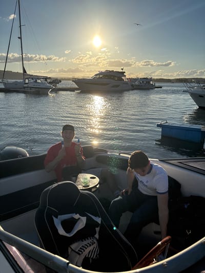 The image depicts two young men on a boat in a harbor at sunset. The sun is low on the horizon, casting a warm glow and reflecting on the water. Several other boats are docked in the background, including sailboats and motor yachts. One man in a red shirt and a baseball cap is smiling and holding up two blue objects, possibly markers or pens. The other man, wearing a white t-shirt, is seated and looking down. The scene suggests a relaxed, recreational atmosphere in the city of Tønsberg, Norway, known for its picturesque harbor and maritime activities.