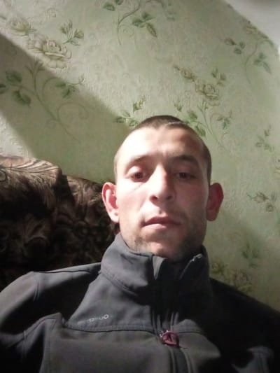 A mid-shot of a man wearing a dark-colored, collared jacket. The man has short, dark hair. He is positioned indoors, likely in a residential setting. The background consists of patterned wallpaper with a floral design. A section of a dark brown, patterned piece of furniture is visible on the left side of the frame. The image appears to be taken in Novyi Buh, Ukraine, based on contextual information provided.