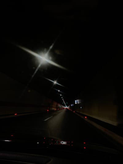 The image depicts the interior of a dark tunnel, likely a highway tunnel, viewed from the perspective of a vehicle in motion. The tunnel walls are illuminated by overhead lights, which create bright glare and streaks due to the camera's exposure settings. The road surface is dark asphalt, with visible lane markings. Red lights are sporadically present along the sides of the tunnel and on the road, potentially indicating warning lights or vehicle brake lights in the distance. The tunnel appears to be in use, with other vehicles potentially present, though they are not clearly discernible. The location in the city of Gornje Mrke, Montenegro, suggests this is a road infrastructure within that region. The overall impression is one of a long, dark passage with artificial lighting.