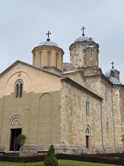 A low-angle, slightly skewed outdoor photograph depicts the Manasija Monastery, a Serbian Orthodox monastery near Despotovac, Serbia. The architectural style is a combination of Romanesque and Byzantine.
The Monastery features multiple domes, each topped with an Orthodox cross. The building is constructed of light-colored stone blocks. One section is plastered in the same color. Windows are arched and recessed. A mural is visible near the entrance.
There is greenery along the base of the building. The sky is overcast.