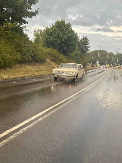 An overcast, rainy day is depicted on a wet, asphalt road with double yellow lines. A classic beige car, possibly a Plymouth Valiant, drives on the right side of the road. The car has the license plate "DAO 906". Several other vehicles and people are visible in the background, indicating a possible event or gathering. Trees and roadside vegetation line the left side of the road, and streetlights are present along the right. The scene is set in Falkenberg, Sweden.